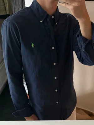 Marinblå skjorta från Ralph Lauren - Klassisk marinblå skjorta från Ralph Lauren med button-down krage och grönt broderat logotyp på bröstet. Skjortan har långa ärmar, vita knappar och är tillverkad i bomull. Perfekt för en clean och stilren look.