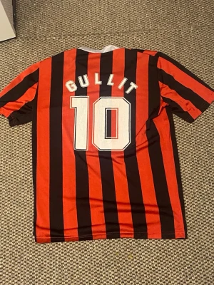 AC Milan retro tröja Gullit #10 - Säljer en retro AC Milan fotbollströja med klassiska röda och svarta vertikala ränder, vit V-ringning och klubbmärke på bröstet. På ryggen står det 'Gullit' och nummer 10 i vitt. Perfekt för dig som älskar vintage fotbollsstil.
