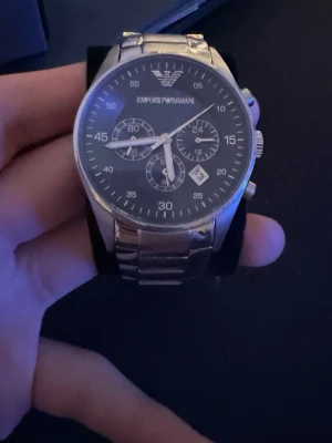 Emporio Armani silverfärgad klocka ( KOM MED BUD) - Stilren klocka från Emporio Armani med svart urtavla och silverfärgat armband i metall. Klockan har tre mindre urtavlor, datumvisning och tydliga vita markeringar. Perfekt accessoar för dig som gillar klassisk och modern design. 2 små repor som man kan se på bild 2