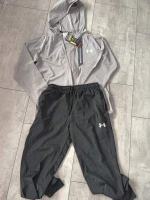Grått träningsset från Under Armour - Snyggt träningsset från Under Armour med en ljusgrå zip-hoodie och mörkgrå byxor. Jackan har huva, dragkedja och fickor med dragkedja samt logga på bröstet och ärmen. Byxorna har resår i midjan och logga på benet. Perfekt för träning eller uppvärmning.