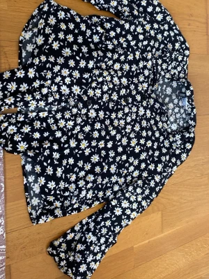 Blommig svart blus från H&M - Supersöt svart blus från H&M med vita och gula prästkragar över hela tyget. Blusen har lång ärm, knappar framtill och en luftig, flowy passform. Perfekt för dig som vill ha en lekfull och trendig look med blommigt mönster.