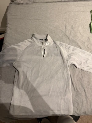 Ljusgrå halvzip-tröja från FootJoy - Ljusgrå sportig halvzip-tröja från FootJoy med hög krage och dragkedja framtill. Tröjan har långa ärmar och är tillverkad i ett mjukt, stretchigt material som passar perfekt för golf eller annan träning. Enkel och stilren design med FJ-logga på kragen.