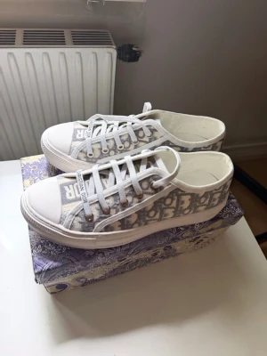 Dior  sneakers  - Snygga låga sneakers från Dior med klassiskt grått och vitt Oblique-mönster. Skorna har vita snören, gummisula och detaljer i canvasmaterial. Perfekta för dig som vill ha en trendig och lyxig look med tydlig logga på tungan och sidan.