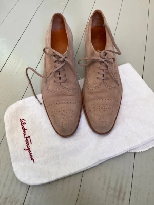 Salvatore Ferragamo skor - Snygga beige loafers i äkta mocka från Salvatore Ferragamo med klassisk broguedetalj på tån och snörning. Skorna har en elegant, smal siluett och bruna lädersulor. Perfekta för dig som gillar stilrena och tidlösa skor.