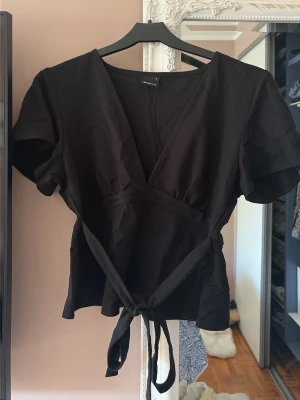 Svart omlottopp med knyt från Gina Tricot - Svart omlottopp från Gina Tricot med korta ärmar och knytband i midjan. Toppen har en djup v-ringning och är tillverkad i mjuk bomull. Perfekt att styla med jeans eller kjol för en trendig look.