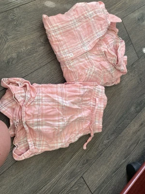 Rosa rutigt pyjamasset i bomull - Mysigt tvådelat pyjamasset i ljusrosa med vitt rutmönster. Setet består av en långärmad skjorta med knappar och bröstficka samt matchande shorts med dragsko i midjan. Perfekt för chillkvällar hemma.