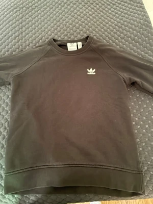 Svart sweatshirt från Adidas - En stilren svart sweatshirt från Adidas med det klassiska vita treklöver-loggan broderad på bröstet. Tröjan har rund halsringning och långa ärmar. Tillverkad i mjukt bomullsmaterial som är skönt mot huden. Perfekt för en avslappnad och sportig look.