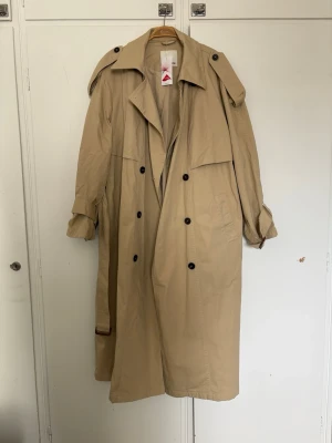 Beige trenchcoat från Bonprix - Stilren beige trenchcoat från Bonprix med klassisk dubbelknäppning, axelklaffar och bälte i midjan. Tillverkad i ett tåligt material med inslag av Lycra för extra komfort. Perfekt oversized look och tidlös design. 6 bilden visar den rätta färgen. 