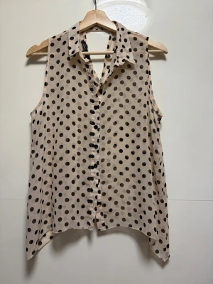 Beige prickig ärmlös blus - Säljer en beige ärmlös blus med svarta prickar och knappar framtill. Blusen har en klassisk krage och asymmetrisk nederkant som ger en cool touch. Perfekt för dig som gillar mönster och vill ha något lätt och luftigt till sommaren.