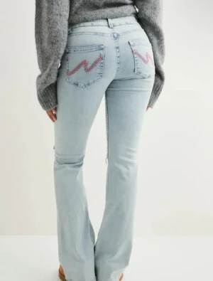 Ljusblå bootcut jeans med slitningar - Säljer ett par ljusblå bootcut jeans med slitna knän och broderade detaljer i rosa på bakfickorna. Jeansen har hög midja och klassisk femficksdesign. Perfekta för en avslappnad och trendig look.
