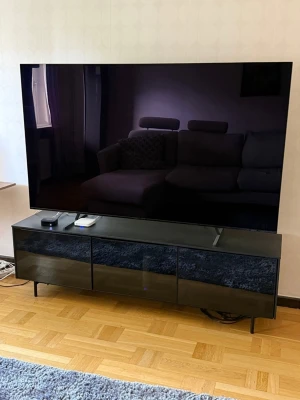 Stor svart platt-TV med TV-bänk - Säljer en stor svart platt-TV med supertunn design och modern look. TV:n står på en stilren svart TV-bänk med tre luckor och höga ben. Perfekt för dig som vill ha en minimalistisk och snygg setup i vardagsrummet. Fjärrkontroll och tillbehör kan placeras på bänken.