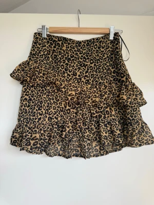 Leopardmönstrad volangkjol - Kort kjol med leopardmönster i beige och svart. Kjolen har volangdetaljer längs nederkanten och sidorna för en extra trendig look.