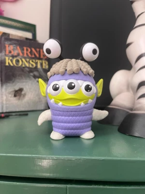 Toy Story alien figur i monsterdräkt - Rolig samlarfigur föreställande en Toy Story alien utklädd till monster med lila kropp, gröna öron, tre ögon och två stora ögon på huvudet. Tillverkad i plast med detaljer som vita tänder och grått hår. 