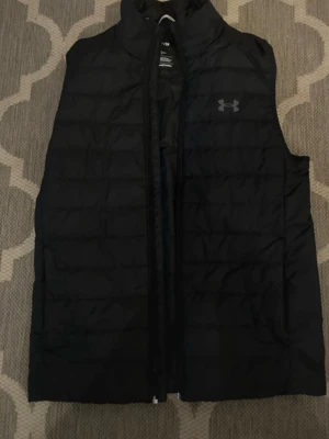 Svart dunväst från Under Armour - Snygg svart dunväst från Under Armour med diskret logga på bröstet. Västen har hög krage, dragkedja framtill och quiltad design som ger en sportig look. Perfekt för lager-på-lager och håller dig varm under kyliga dagar.