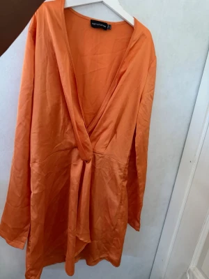 Orange omlottklänning från PLT - Säljer en snygg orange omlottklänning från Nelly med lång ärm och v-ringad hals. Klänningen har knyt i midjan och är tillverkad i ett glansigt material som ger en lyxig känsla. Perfekt för dig som vill sticka ut med färg!