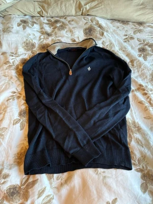 Mörkblå stickad tröja från Morris - Mörkblå stickad tröja från Morris i 100% merinoull. Tröjan har halv dragkedja, beige insida på kragen och en liten broderad logga på bröstet. Perfekt för dig som gillar klassisk och stilren design.