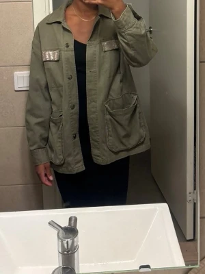 Olivgrön overshirt med fickor - Stilren olivgrön overshirt med stora framfickor och dekorativa detaljer i form av glittriga band på bröstfickorna. Rak passform och knäppning framtill. Perfekt för lager på lager och enkel att styla till olika outfits.