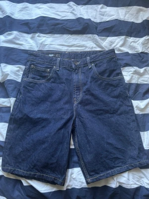 Mörkblå denimshorts från Lee - Raw denim mörkblå denimshorts från Pull & bear med fem fickor och kontrastsömmar. Modellen har normal passform och bälteshällor i midjan. Perfekta för sommaren.