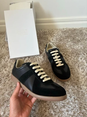 Svarta sneakers från Maison Margiela - Säljer ett par svarta sneakers från Maison Margiela med beige sula och vita snören. Skorna har en mix av mocka och läderdetaljer, samt klassisk låg silhuett. Perfekta för dig som gillar stilrena och tidlösa sneakers.