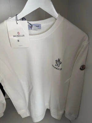 Vit sweatshirt från Moncler - Snygg vit sweatshirt från Moncler med rund halsringning och diskret logga broderad på bröstet samt patch på ärmen. Tillverkad i mjukt material som känns skönt mot huden. Perfekt för en clean och stilren look.