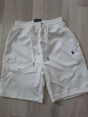 Vita shorts från Polo Ralph Lauren - Säljer ett par vita shorts från Polo Ralph Lauren med elastisk midja och snörning. Klassisk logga broderad på vänster ben. Sköna och stilrena, perfekta till sommaren. Två sidofickor och mjukt material.