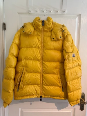 Gul dunjacka från Moncler - Snygg gul dunjacka från Moncler med avtagbar huva och två framfickor med dragkedja. Jackan har en patch med Moncler-logga på ärmen och quiltad design som ger en puffig look. Perfekt för kalla vinterdagar och riktigt trendig färg. Finns några små fläckar se bild 5 har inte försökt tvätta bort de men tror inte det ska vara några problem och få bort de i tvätten. 