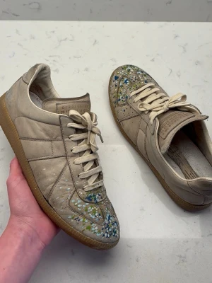 Maison Margiela sneakers med färgstänk - Beiga sneakers från Maison Margiela i läder med unika färgstänk i blått, grönt och vitt på tån. Klassisk låg modell med beige snörning och gummisula. Perfekt för dig som vill sticka ut med en kreativ och trendig look.