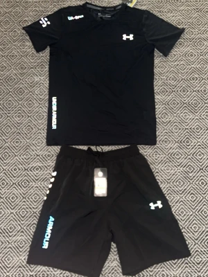 Svart träningsset från Under Armour - Snyggt svart träningsset från Under Armour med t-shirt och shorts. Setet har vita och turkosa loggor och textdetaljer. Tillverkat i lätt och snabbtorkande material som passar perfekt för träning och sportaktiviteter. T-shirten har rund hals och shortsen har dragsko i midjan. Storlek M men passar också S