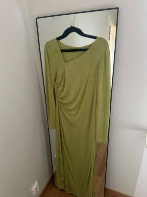 Grön långklänning med drapering - Snygg grön långklänning med asymmetrisk halsringning och draperad detalj framtill. Klänningen har långa ärmar och är tillverkad i ett mjukt, stretchigt material som ger en elegant siluett. Perfekt för dig som vill sticka ut med en unik look.