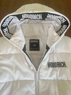 Vit dunjacka med huva från Hoodrich - Snygg vit dunjacka från Hoodrich med huva och dragkedja framtill. Jackan har svart Hoodrich-logga på bröstet och tryck på insidan av huvan. Två fickor framtill och quiltad design för extra värme. Perfekt för kalla vinterdagar. Den är XS MEN PASSAR MER SOM M