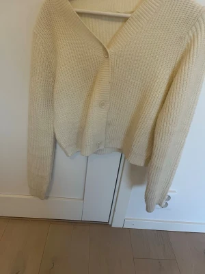 Krämvit stickad kofta från KappAhl - Mysig krämvit stickad kofta från KappAhl med v-ringning och stora knappar framtill. Koftan har lång ärm och ribbade muddar, perfekt för lager på lager. Passar dig som gillar en enkel och stilren look.