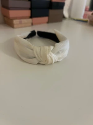 Vitt knutdiadem i tyg - Snyggt vitt diadem med knut framtill. Tillverkat i mjukt tyg med en bred form som ger en trendig look. Insidan är svart för extra kontrast och komfort. Perfekt accessoar för att lyfta din outfit.