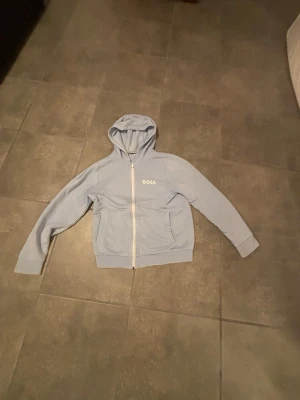 Ljusblå hoodie från BOSS med dragkedja - Ljusblå hoodie från BOSS med vit dragkedja, huva och två fickor framtill. Tillverkad i mjukt bomullstyg och har ribbade muddar vid ärmsluten. BOSS-logga broderad på bröstet för en clean look.