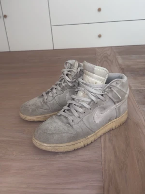 Nike Dunk High grå mockasneakers - Snygga Nike Dunk High sneakers i grå mocka med vita detaljer och klassisk swoosh på sidan. Skorna har högt skaft, perforerad tå och beige sula för en retrovibe. Perfekta för dig som gillar streetwear och vill ha en stilren look.