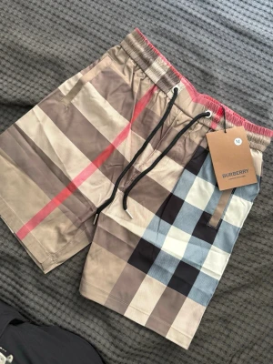 Rutig Burberry shorts med snörning - Snygga shorts från Burberry i klassiskt rutigt mönster med beige, svart, vitt och röda detaljer. Shortsen har elastisk midja med snörning och bakficka. Perfekta för sommaren och ger en trendig look.