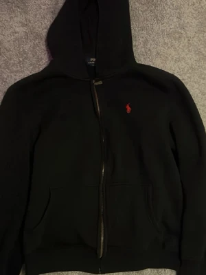 Svart hoodie från Polo Ralph Lauren - Svart hoodie med dragkedja från Polo Ralph Lauren. Klassisk design med röd broderad logga på bröstet och två fickor framtill. Tillverkad i mjukt material och har en stor huva för extra komfort. Vet ej storleken men passar en som är runt 14,16 år, 170cm.. storlek M funkar som S 