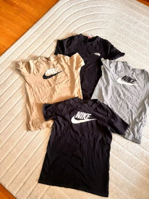 Nike och The North Face t-shirts - Fyra snygga t-shirts från Nike och The North Face. En svart och en beige Nike t-shirt med stor logga, en grå Nike t-shirt med logga och en svart The North Face t-shirt. Alla har klassisk passform och är kortärmade, perfekta till vardags. Materialet känns som mjuk bomull.