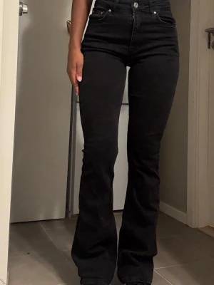 Svarta bootcut jeans med hög midja - Snygga svarta bootcut jeans med mid midja och klassiska fem fickor. Jeansen har en figurnära passform upptill och utsvängda ben nertill. Perfekta för dig som gillar en trendig och stilren look.