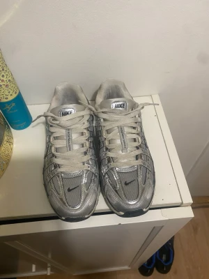 Nike silverfärgade sneakers - Säljer ett par Nike sneakers i silver och grått med mesh och syntetmaterial. Skorna har klassisk snörning, reflekterande detaljer och den ikoniska swoosh-loggan på sidan och framtill. Perfekta för dig som gillar sportig och trendig stil.