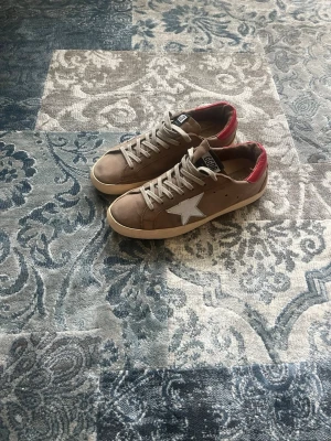 Beige Golden Goose sneakers med röd häl storlek 41 - Snygga beige sneakers från Golden goose med vit stjärndetalj på sidan och röd häl. Skorna har beige snörning och en klassisk låg siluett. Ovandelen är i mocka och sulan är vit. Perfekta för dig som vill ha en unik touch på din outfit. Skorna har storlek 41