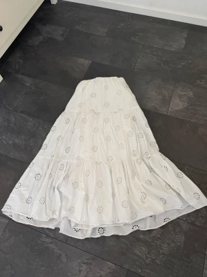 Vit långkjol med broderade detaljer - Säljer en vit långkjol med broderade blommönster och volang. Kjolen har en luftig och flowy passform, perfekt för varma dagar. Resår i midjan för extra komfort och snygg siluett.