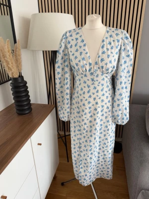 Blommig långklänning med v-ringning Gina Tricot 38 - Långklänning med v-ringad front och puffiga långärmar. Klänningen är vit med blått blommigt mönster och har en figurnära passform. Perfekt för sommarens alla tillfällen. Materialet ser ut att vara bomull eller linne för en luftig känsla.