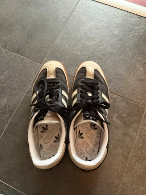Adidas Samba svarta sneakers - Klassiska Adidas Samba sneakers i svart med vita detaljer och bruna gummisulor. Skorna har de ikoniska tre ränderna på sidan och svart snörning. Ovandelen är i mocka och läder, vilket ger en retro och sportig vibe. Perfekta för dig som gillar streetwear och tidlös stil.
