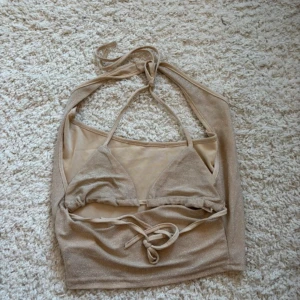 Beige glittrig bikini med snörning - Säljer en beige bikini med glittrigt tyg och snygg snörning både i nacken och runt midjan. Bikinin har trekantsformade kupor och ett halterneck-band. Perfekt för dig som vill ha en trendig och unik look på stranden!