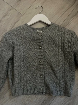 Grå kabelstickad kofta från H&M - Mysig grå kabelstickad kofta från H&M med rund halsringning och dekorativa pärlknappar