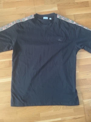 Svart t-shirt med rutiga axelband - Snygg svart t-shirt från Burberry med klassiska rutiga band längs axlarna i beige, rött och svart. T-shirten har korta ärmar och en liten broderad logga på bröstet. Tillverkad i mjuk bomull för skön känsla.