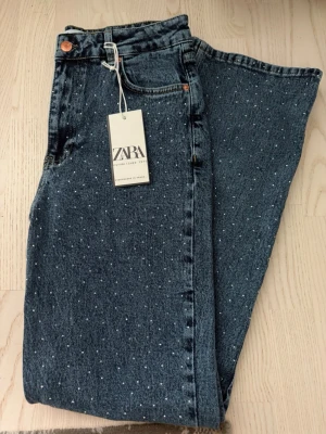 Blå jeans med pärldetaljer från Zara - Snygga blå jeans från Zara med raka ben och små vita pärldetaljer över hela tyget. Klassisk femficksmodell med hög midja och knappgylf. Jeansen är gjorda i kraftig denim och har en trendig look som sticker ut.