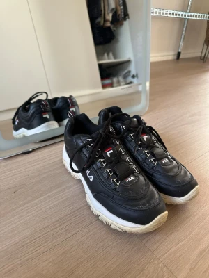 Svarta Fila Disruptor sneakers - Chunky svarta Fila Disruptor sneakers med vit, grov sula och klassisk logga på sidan, plös och häl. Skorna har svarta snören och detaljer i vitt och rött. Tillverkade i syntetmaterial och textil, med låg profil och sportig vibe.