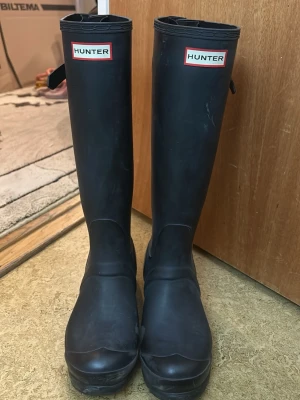 Svarta höga gummistövlar från Hunter - Klassiska svarta höga gummistövlar från Hunter med logotyp framtill. Stövlarna har rund tå, justerbar rem upptill och är tillverkade i slitstarkt gummi. Perfekta för regniga dagar och festivaler.
