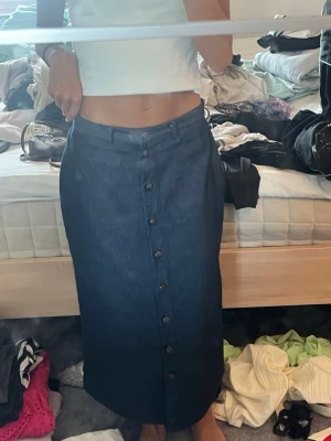 Mörkblå långkjol i denim med knappar - Säljer en mörkblå långkjol i denim med knappar hela vägen framtill. Kjolen har hög midja och en rak passform som ger en clean och stilren look. Perfekt att matcha med crop tops eller stickade tröjor.